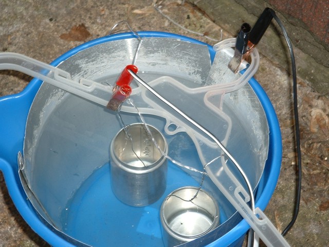 The shakers hanging from their wires in a plastic bucket. A red (positive) clamp from a car battery charger is attached to the lead wires, while a black (negative) clamp is attached to a large sheet of alumunim lining the bucket.